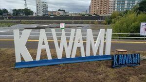 豊平川沿いに設置された白いKAWAMIの大きな文字モニュメント。背後に川と対岸の河川敷が見える