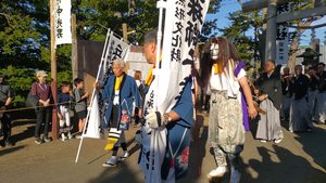 丘珠神社の境内を進む、白い面と長い髪の装束を着けた人物、奉納旗を持つ人たちとお囃子の一行