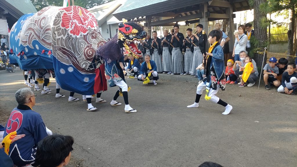 丘珠神社の境内で、大きな獅子と青い法被を着た獅子取りの子どもが向かい合って舞う様子