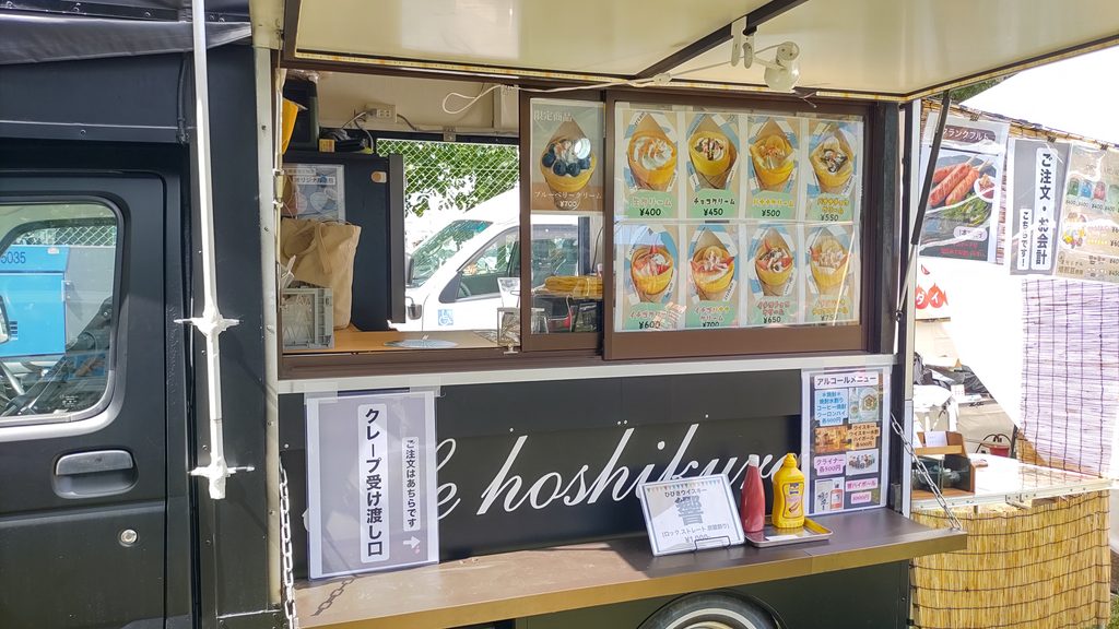 クレープの写真付きメニューが並ぶ黒いキッチンカー。受け渡し口と複数種類のクレープが掲示されている